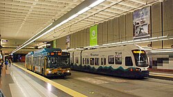 A green bus pulling up to the curb of an underground station, pictured alongside a white train heading in the opposite direction